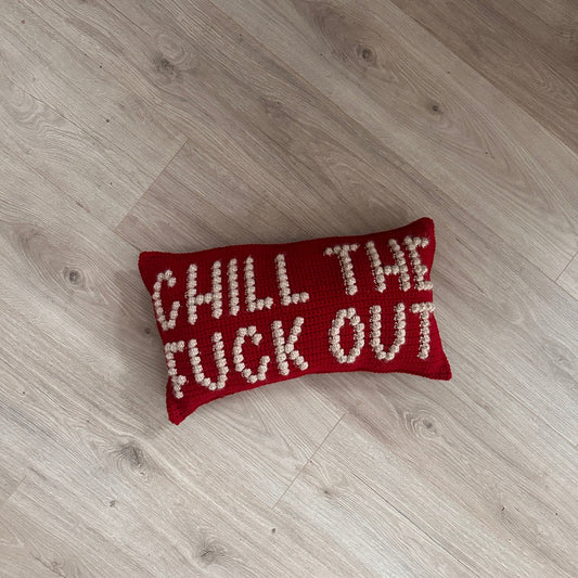 A red crochet pillow with cream colored puffy letters that say &quot;chill the fuck out&quot; lays on a light wood floor.